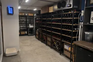 Converted garage with metal racks holding wine bottles, wall-mounted thermometer, humidifier tray in corner, illuminated by cool LED lighting.