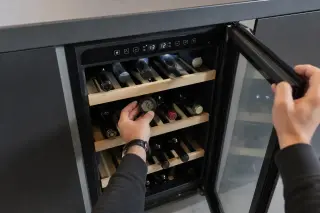 Electric wine cabinet partially open under a contemporary worktop, with a hand checking the door seal and an interior hygrometer visible among stored bottles.