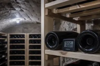 Close-up of a digital hygrometer on a wooden wine rack shelf between two bottle necks in a stone vaulted cellar, with cool LED lighting and rough masonry visible.