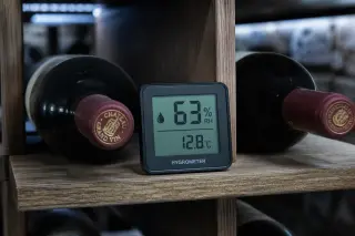 Digital hygrometer between two lying wine bottles on a wooden rack, in a stone cellar under cool LED lighting.