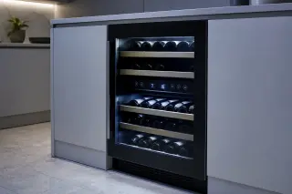 Built-in wine cabinet under a modern kitchen counter, bottles stored horizontally, digital control panel visible and illuminated by cool LED lighting.