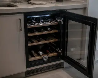 Built-in wine cabinet under a modern kitchen counter, LED-lit interior with bottles on wooden racks and a digital hygrometer, focus on technical storage solution